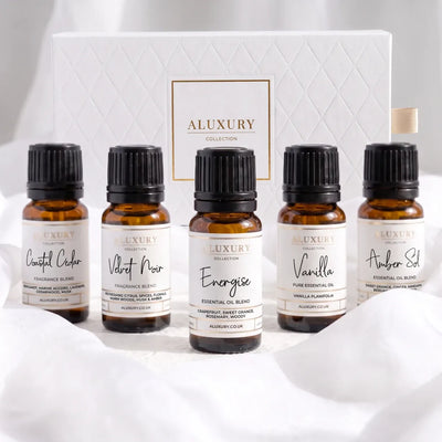 Five amber glass essential oil bottles with black caps from the ALUXURY COLLECTION are arranged in front of a white quilted gift box. The labels read: Central Cider, Velvet Noir, Energise, Vanilla, and Amber St.
