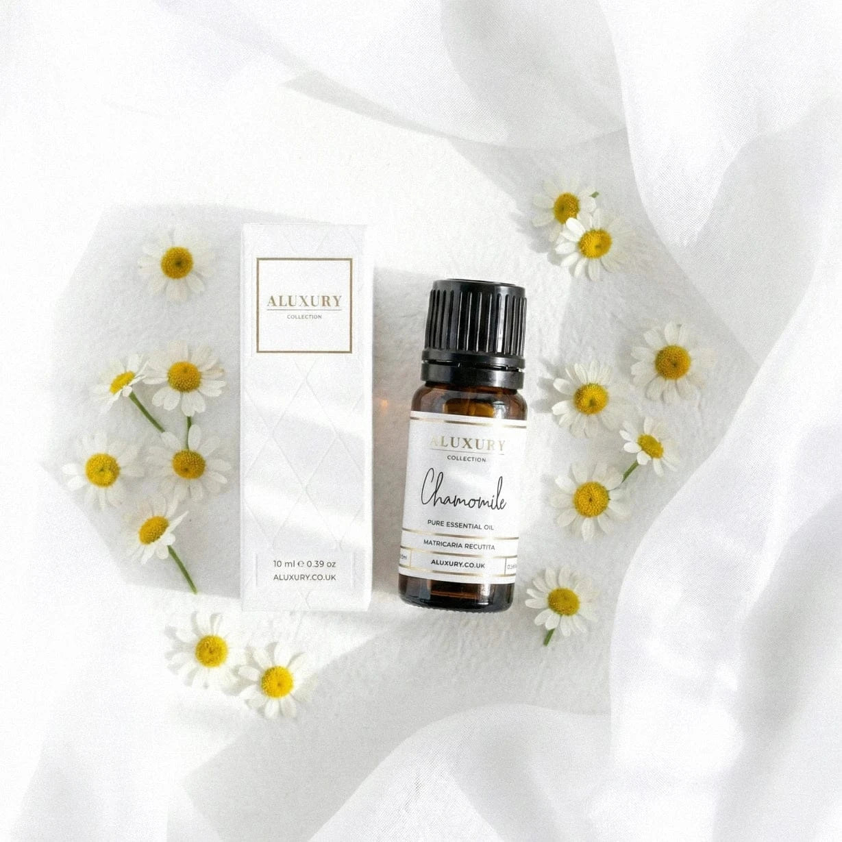 Aluxury chamomile essential oil bottle and packaging on a white fabric background with daisies.