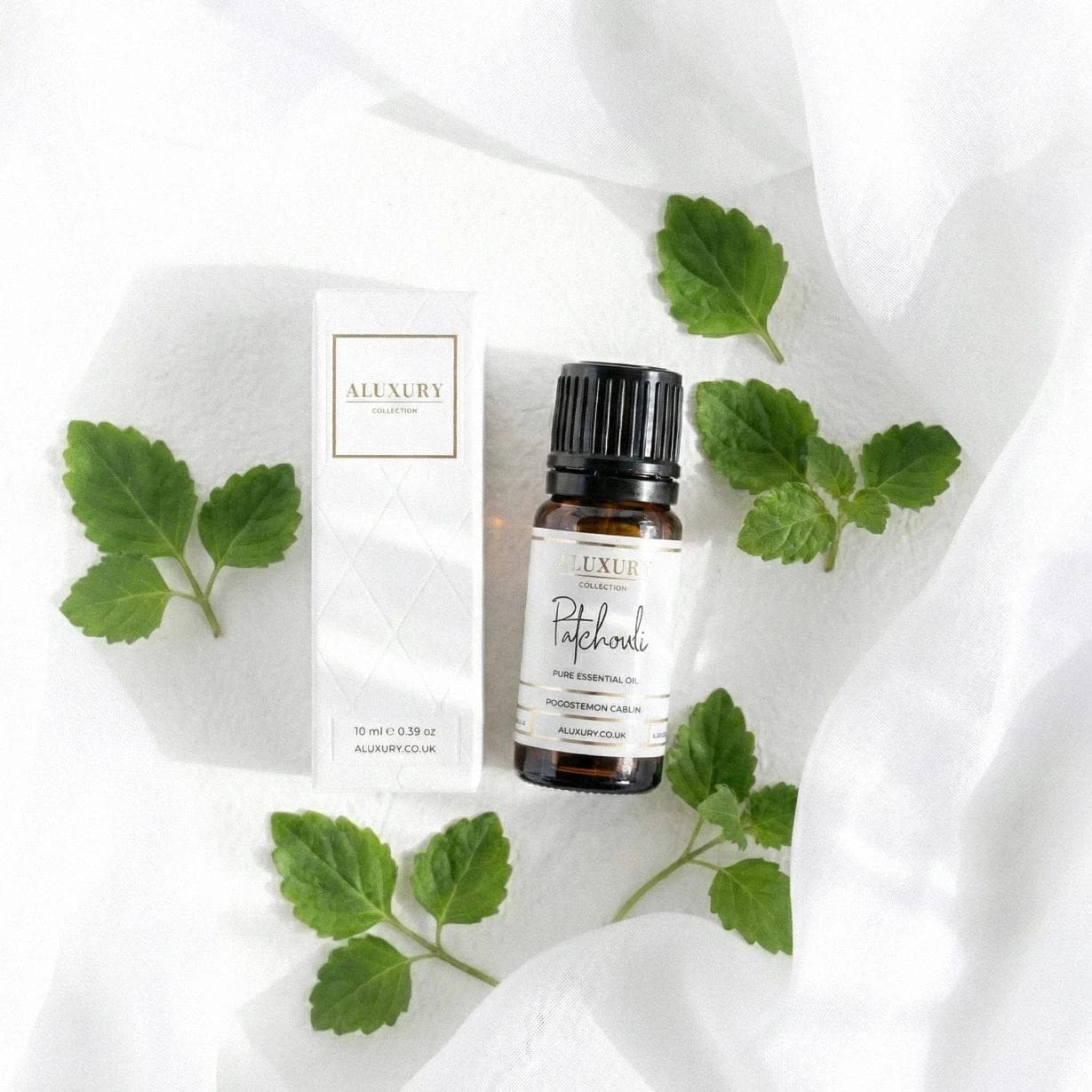 Aluxury patchouli essential oil bottle with green leaves and packaging on a white fabric background