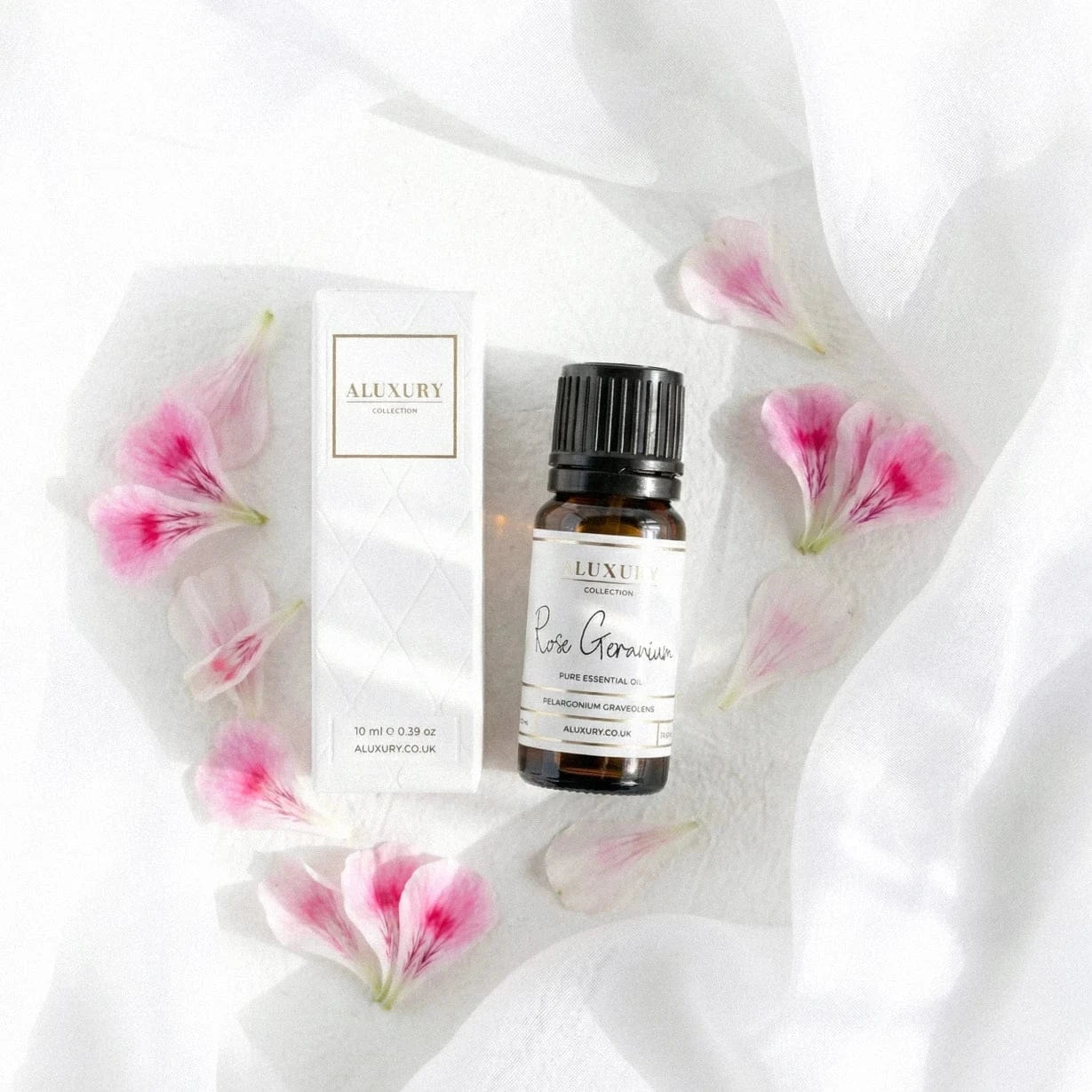 Luxury rose geranium essential oil bottle and packaging on a white fabric background with pink flower petals.