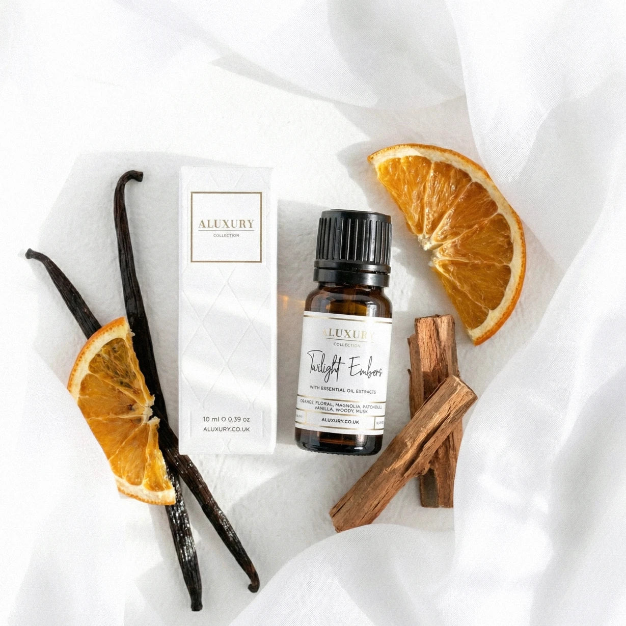 Bottle of 'Twilight Embers' essential oil with vanilla beans, orange slices, and cinnamon sticks on a white fabric background.