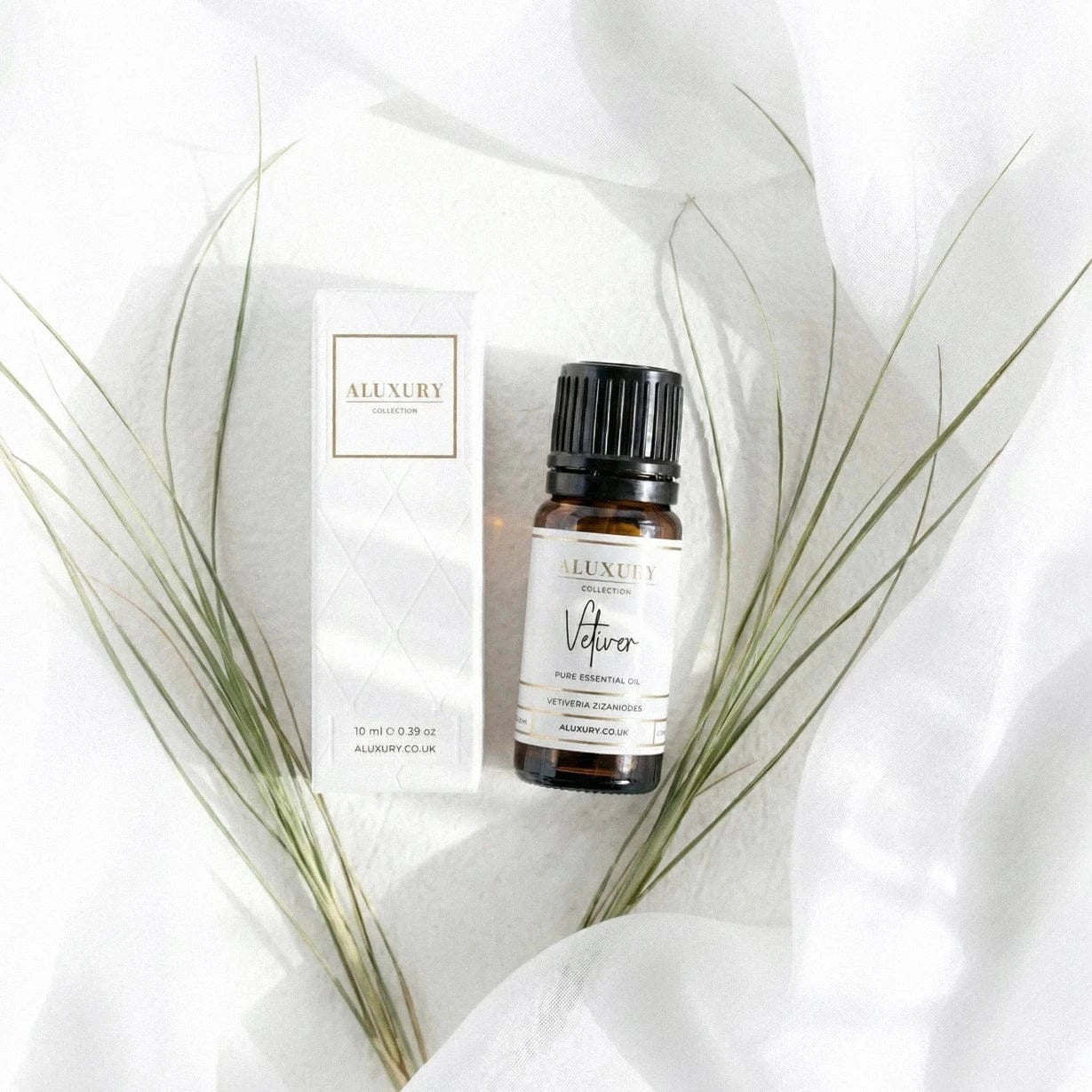 Aluxury Vetiver essential oil bottle and packaging on a white fabric background with green leaves.